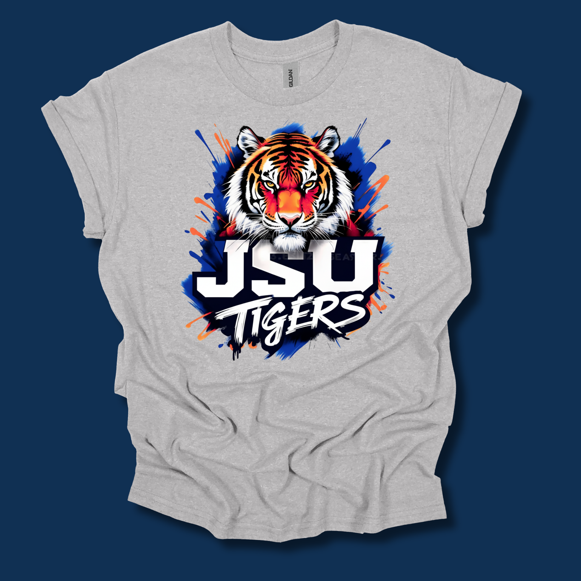 A light heather gray t-shirt with a graphic of a tiger head and the text 'JSU TIGERS' in white, black, and blue paint splatter design, shown against a dark blue background.