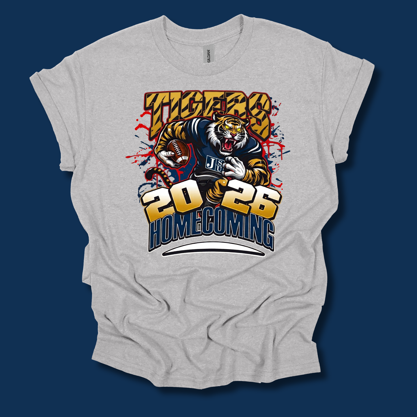 A light heather gray t-shirt with a graphic print of a tiger mascot wearing a blue and gold jersey, with the text 'TIGERS 2026 HOMECOMING' in bold yellow and blue lettering. The t-shirt is displayed flat against a dark navy blue background.
