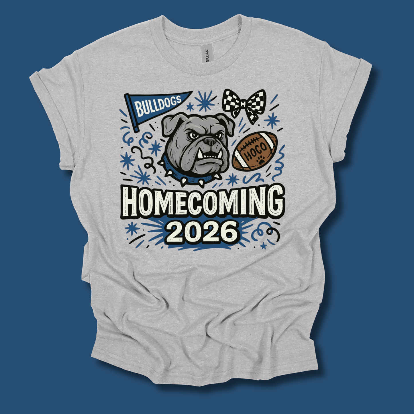 A light heather gray t-shirt with a graphic print for 'Bulldogs Homecoming 2026'. The graphic features a bulldog mascot, a football, a bow tie, and decorative blue stars and stripes.