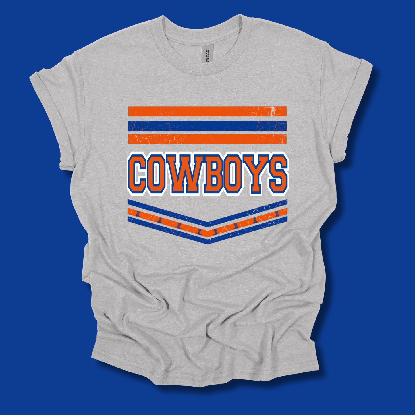 A light heather gray t-shirt with a distressed graphic featuring the word 'COWBOYS' in orange and blue, framed by horizontal orange and blue stripes, displayed on a solid blue background.