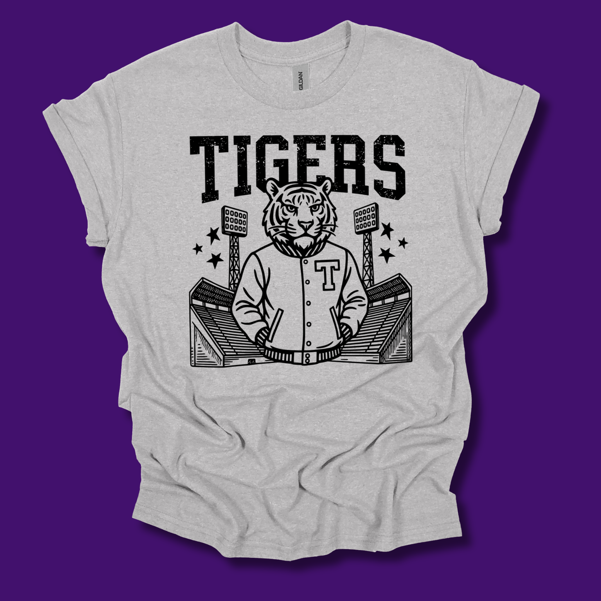 A light heather gray t-shirt with a black graphic print of a tiger mascot wearing a varsity jacket in front of a stadium, with the word 'TIGERS' printed above. The t-shirt is displayed flat against a solid purple background.