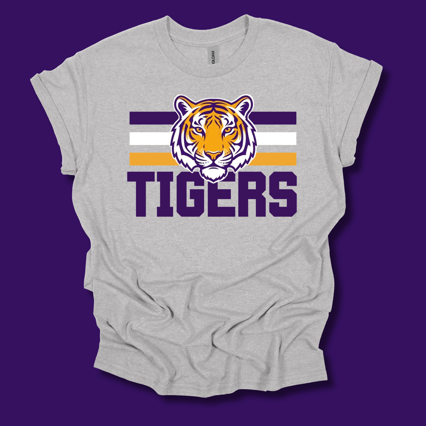 A light heather gray t-shirt with a graphic of a tiger head over purple and white stripes, with the word 'TIGERS' printed below in purple. The t-shirt is displayed flat against a solid purple background.