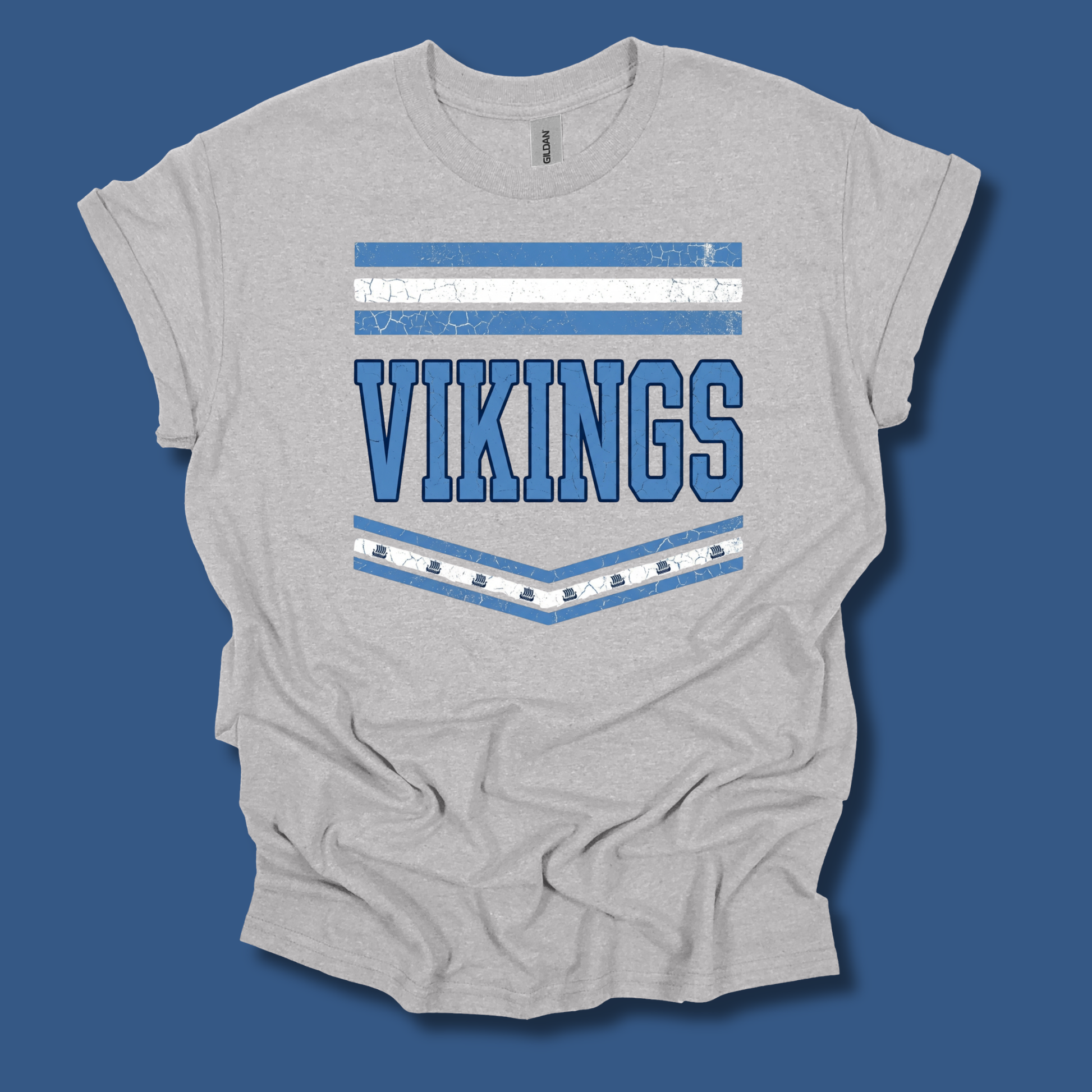 A light heather gray t-shirt with a distressed blue and white graphic featuring the word 'VIKINGS' and horizontal stripes, displayed on a solid blue background.