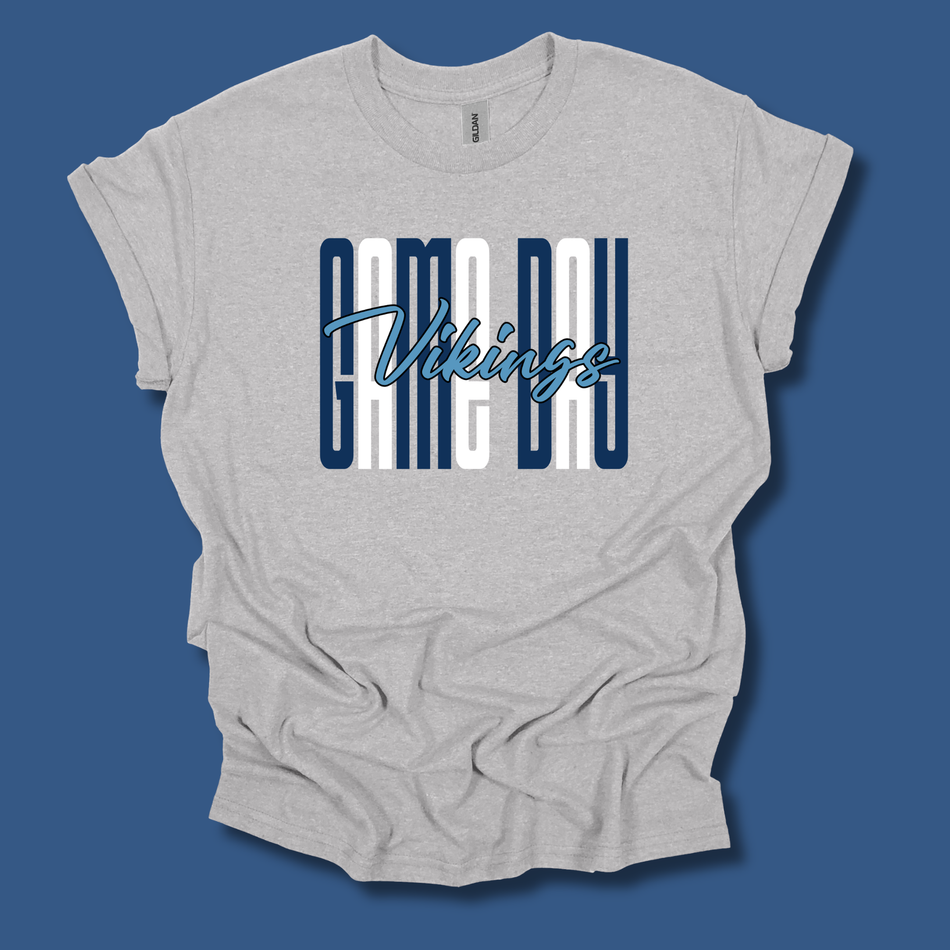 A light heather gray t-shirt with a graphic on the front reading 'GAME DAY Vikings' in navy blue and white, displayed against a solid blue background.