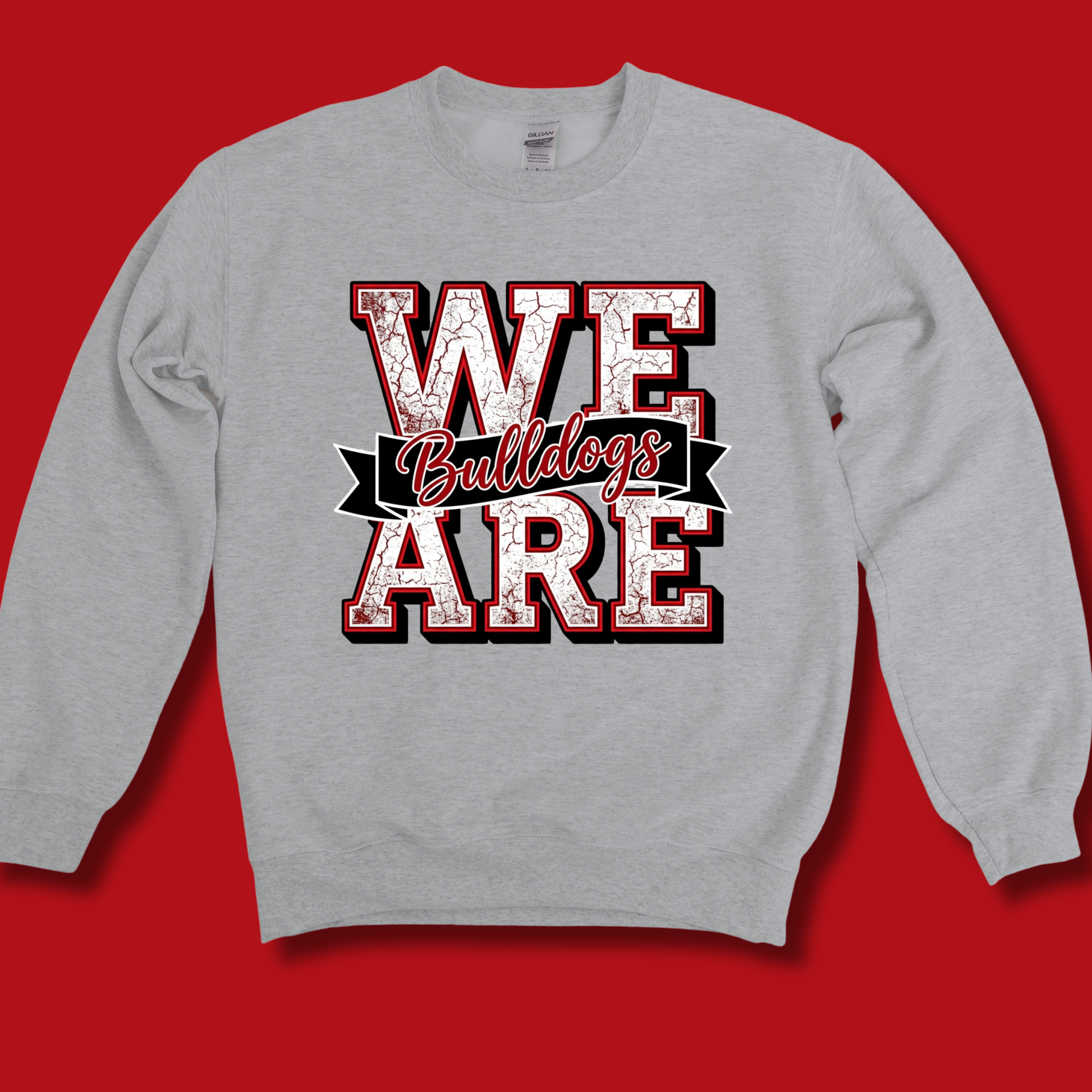 A light gray crewneck sweatshirt with a graphic print reading 'WE Bulldogs ARE' in distressed red, white, and black lettering, displayed against a solid red background.