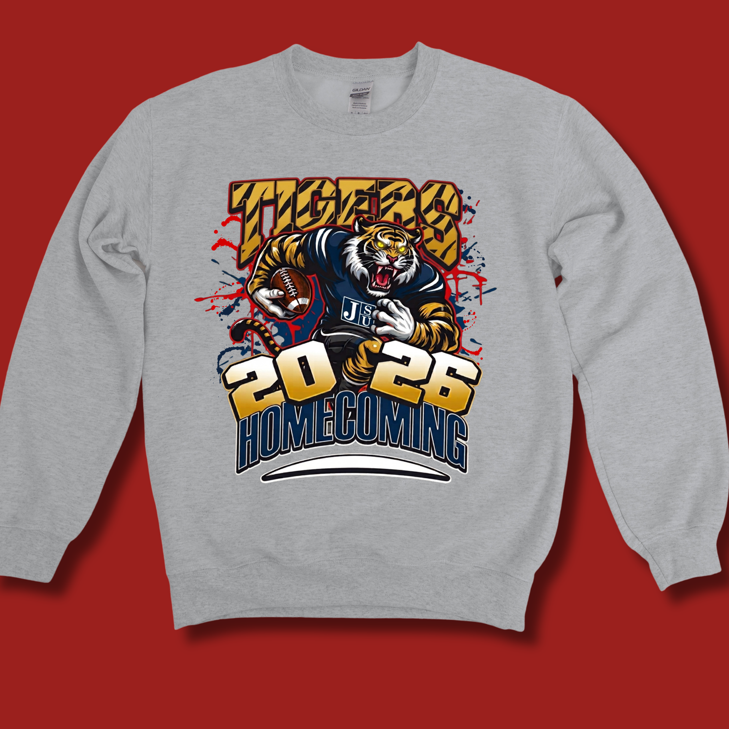 A light gray crewneck sweatshirt featuring a large graphic print of a tiger mascot wearing a navy and gold football jersey, with the text 'TIGERS 2026 HOMECOMING' in bold yellow and blue lettering, displayed against a solid red background.