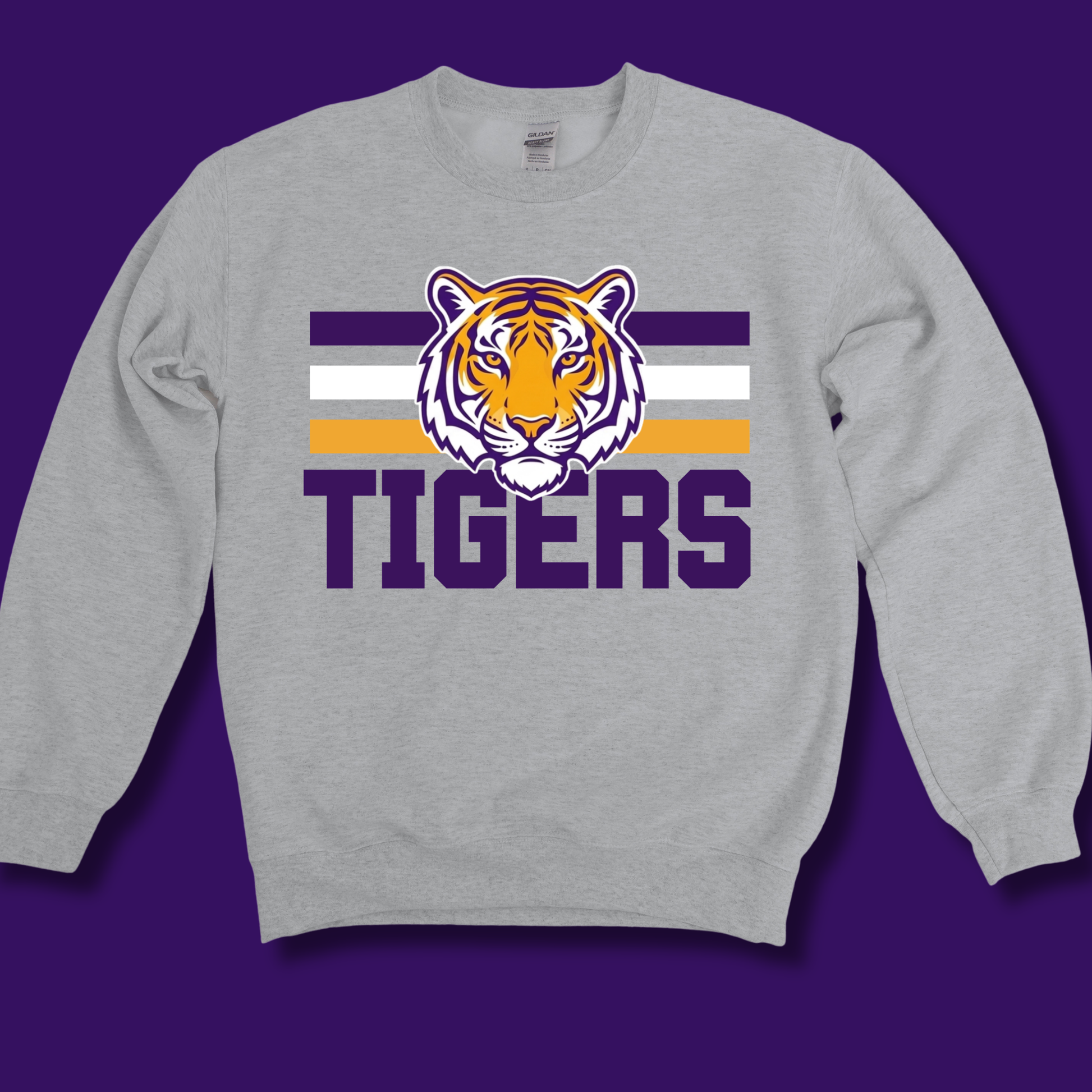 A heather gray crewneck sweatshirt featuring a purple, white, and orange tiger mascot graphic above the word 'TIGERS' in purple, displayed against a solid purple background.
