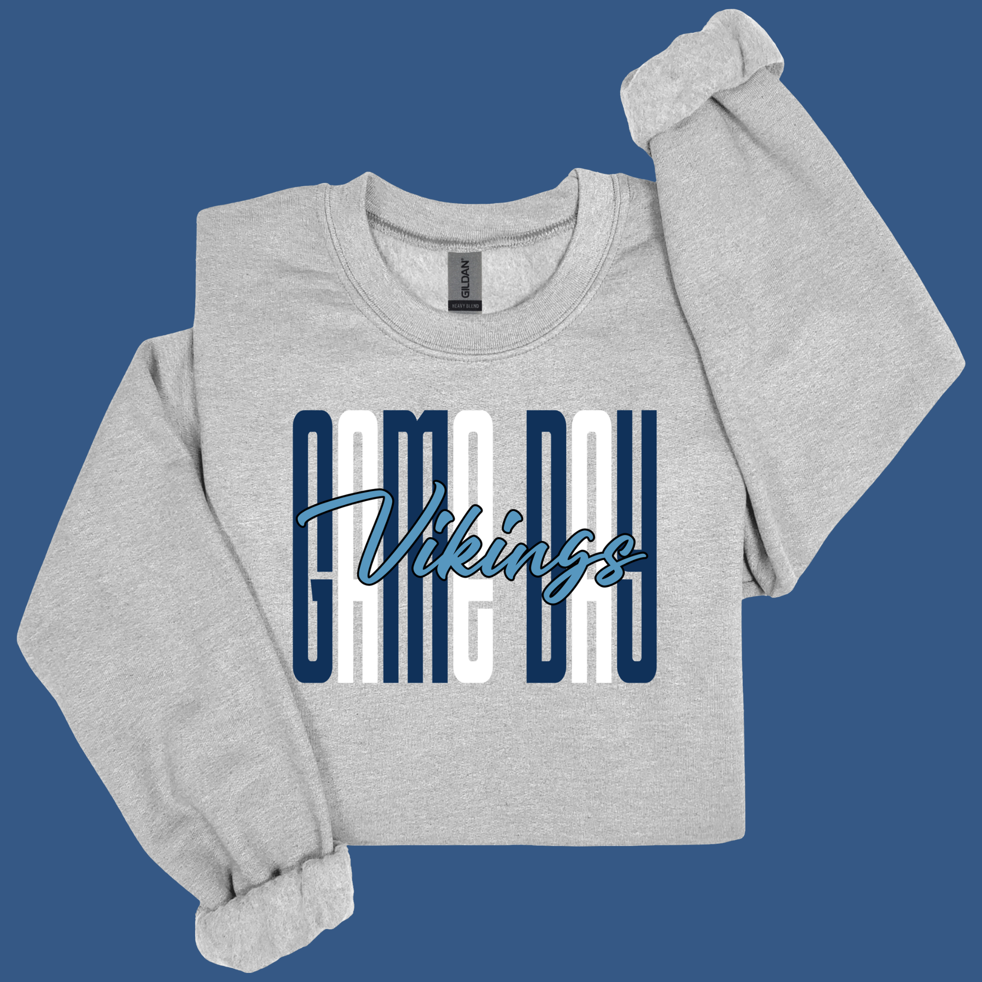 A light heather gray crewneck sweatshirt with rolled cuffs, featuring a graphic print on the front that reads 'GAME DAY Vikings' in navy blue and white. The sweatshirt is displayed flat against a solid blue background.