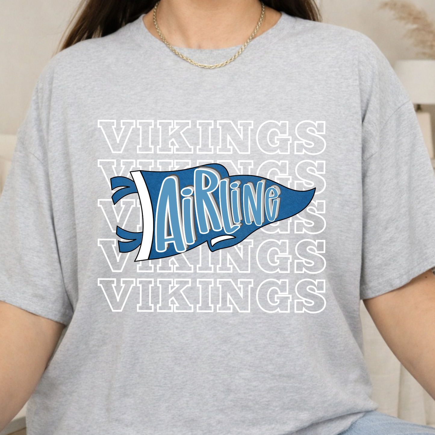 A person wearing a light heather gray t-shirt with a blue and white 'AiRline Vikings' graphic on the front, layered over a repeating white 'VIKINGS' text pattern.