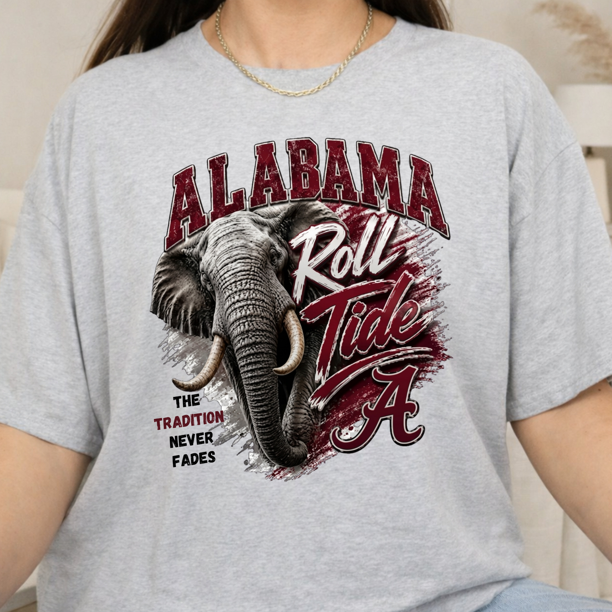 A person wearing a light heather gray graphic T-shirt featuring an elephant and the words 'ALABAMA Roll Tide' in maroon and white, with 'The Tradition Never Fades' printed below.