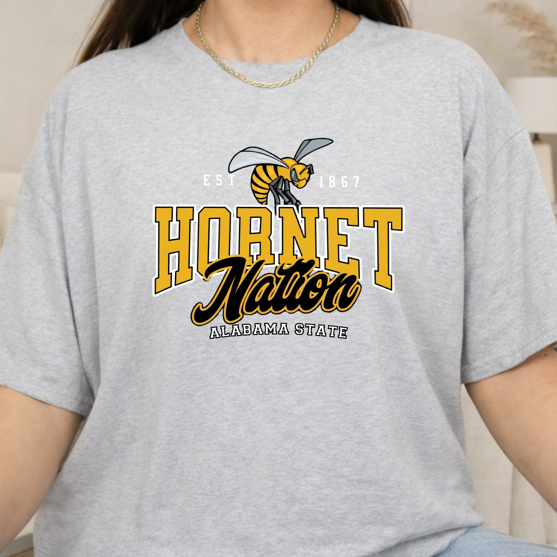 A person wearing a light heather gray short-sleeve t-shirt with a yellow and black 'Hornet Nation Alabama State' graphic on the front.