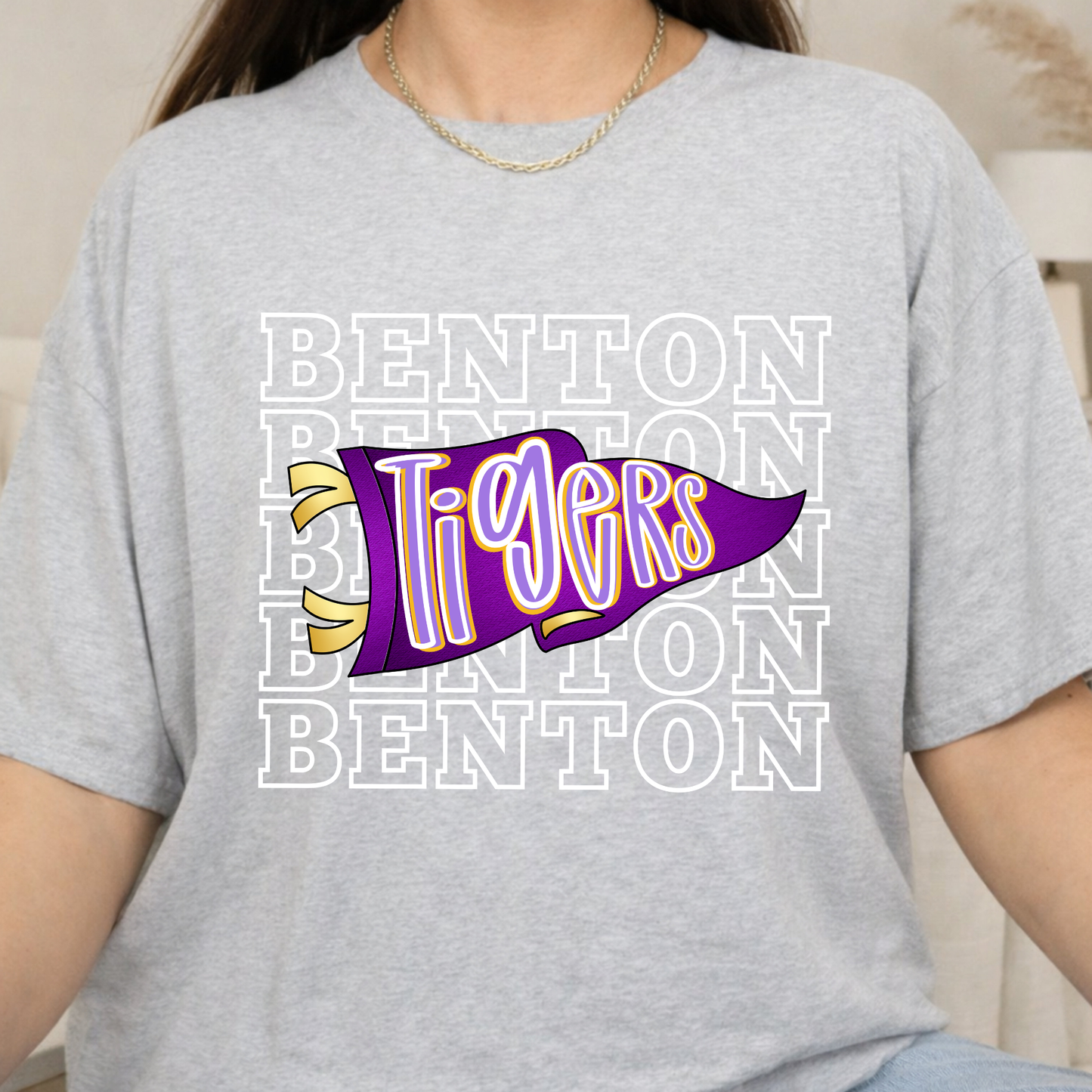 A person wearing a light heather gray t-shirt with a purple and yellow 'Tigers' pennant graphic over a white 'BENTON' text pattern.