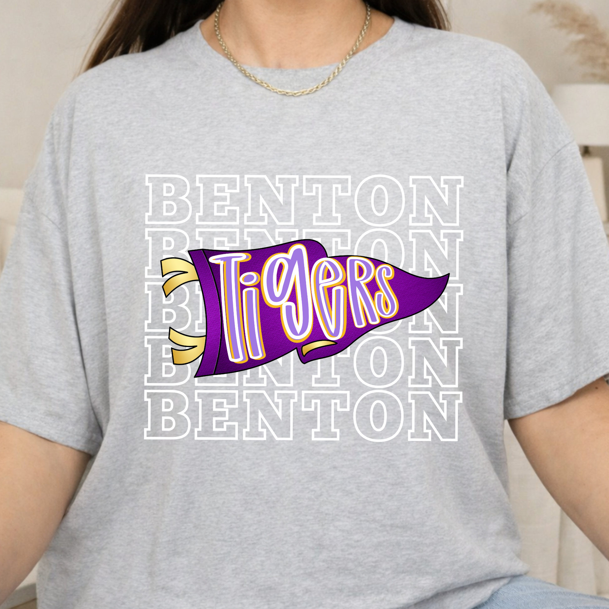 A person wearing a light heather gray t-shirt with a purple and yellow 'Tigers' pennant graphic over a white 'BENTON' text pattern.