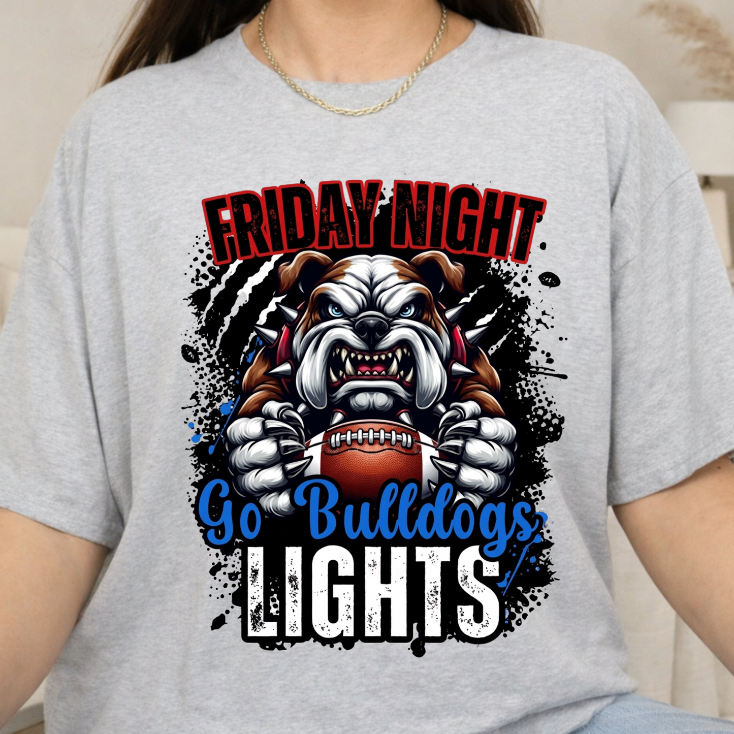 A person wearing a light heather gray graphic T-shirt. The shirt features a bulldog mascot holding a football with the text 'FRIDAY NIGHT Go Bulldogs LIGHTS' in red, blue, and white lettering over black paint splatter graphics.