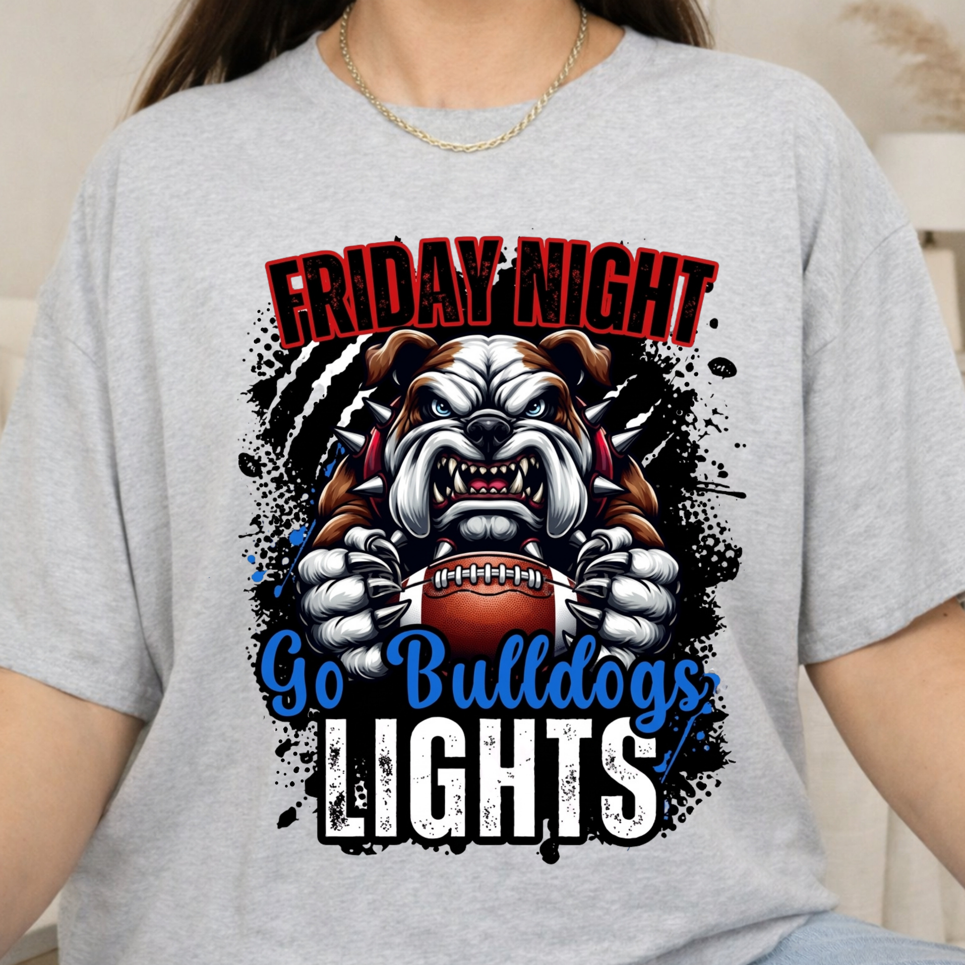 A person wearing a light heather gray graphic T-shirt. The shirt features a bulldog mascot holding a football with the text 'FRIDAY NIGHT Go Bulldogs LIGHTS' in red, blue, and white lettering over black paint splatter graphics.