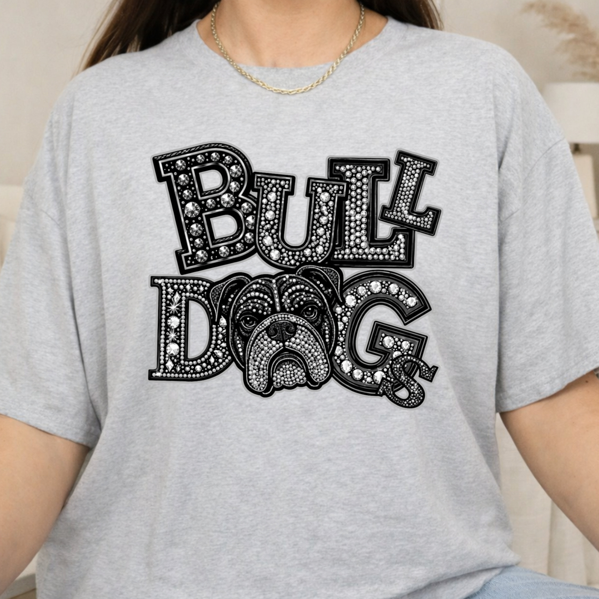 A person wearing a light heather gray t-shirt with a black and white graphic that reads 'BULLDOG' in decorative block letters, where the 'O' is shaped like a bulldog's face. The person is also wearing a gold chain necklace.