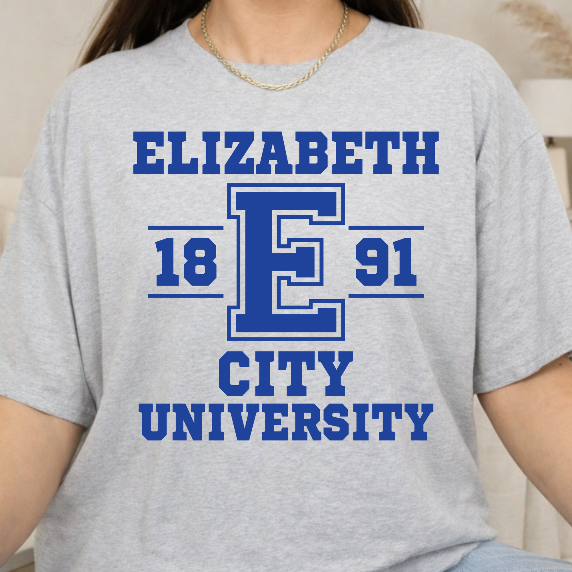 A person wearing a light heather gray short-sleeve T-shirt with a bold blue graphic reading "ELIZABETH E 1891 CITY UNIVERSITY" on the front.