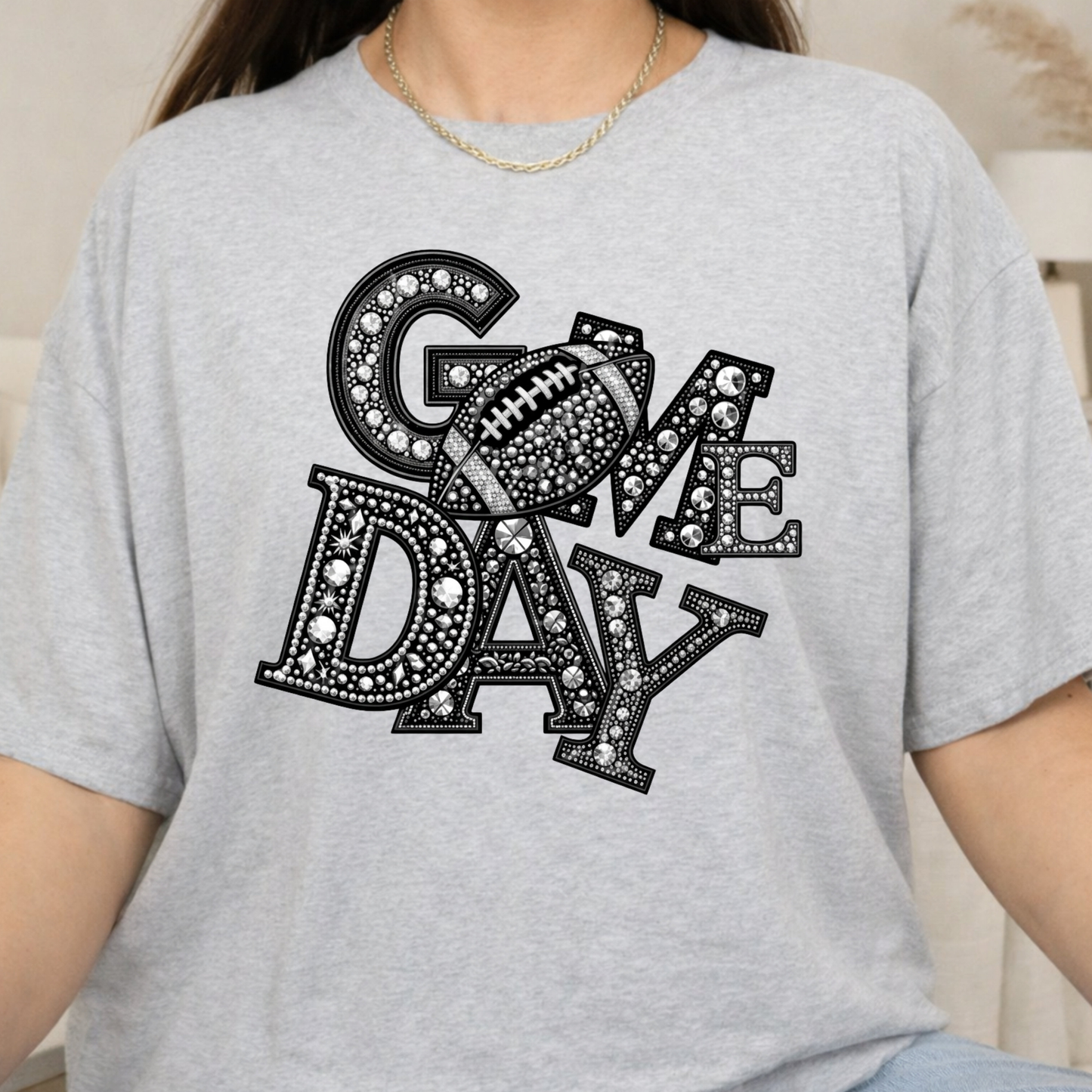A person wearing a light heather gray t-shirt with a black and white rhinestone-style graphic that reads 'GOME DAY' with a football motif, photographed in a soft indoor setting.