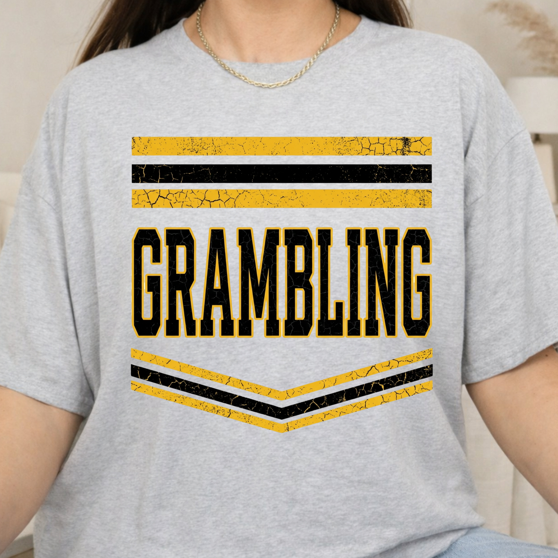 A person wearing a light heather gray t-shirt with a distressed yellow and black graphic that reads 'GRAMBLING' with horizontal stripes above and chevron stripes below.