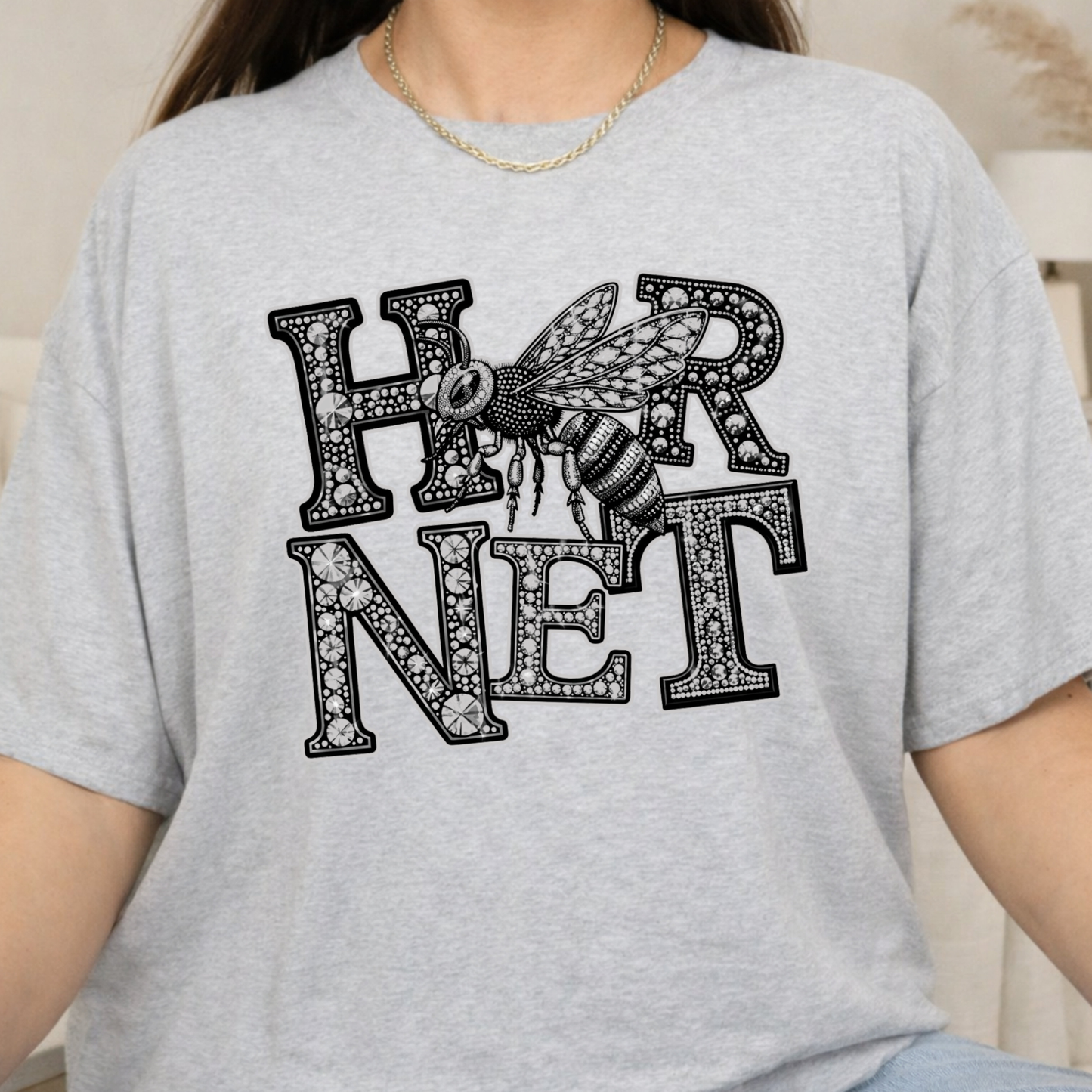 A person wearing a light heather gray t-shirt with a black graphic print that reads 'HARNESS' in decorative block letters with a detailed bee design integrated into the text.