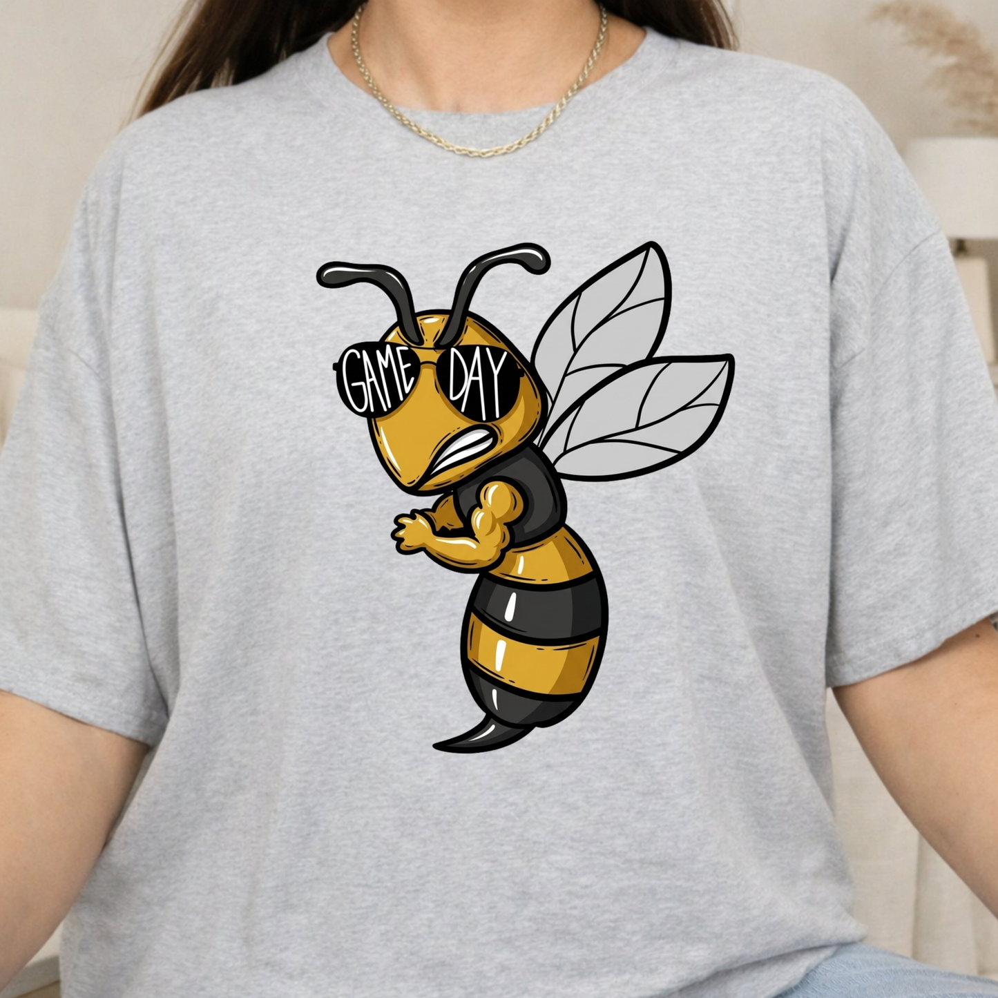A person wearing a light heather gray t-shirt with a cartoon bee graphic on the chest. The bee is yellow and black with the words 'GAME DAY' written on its sunglasses.