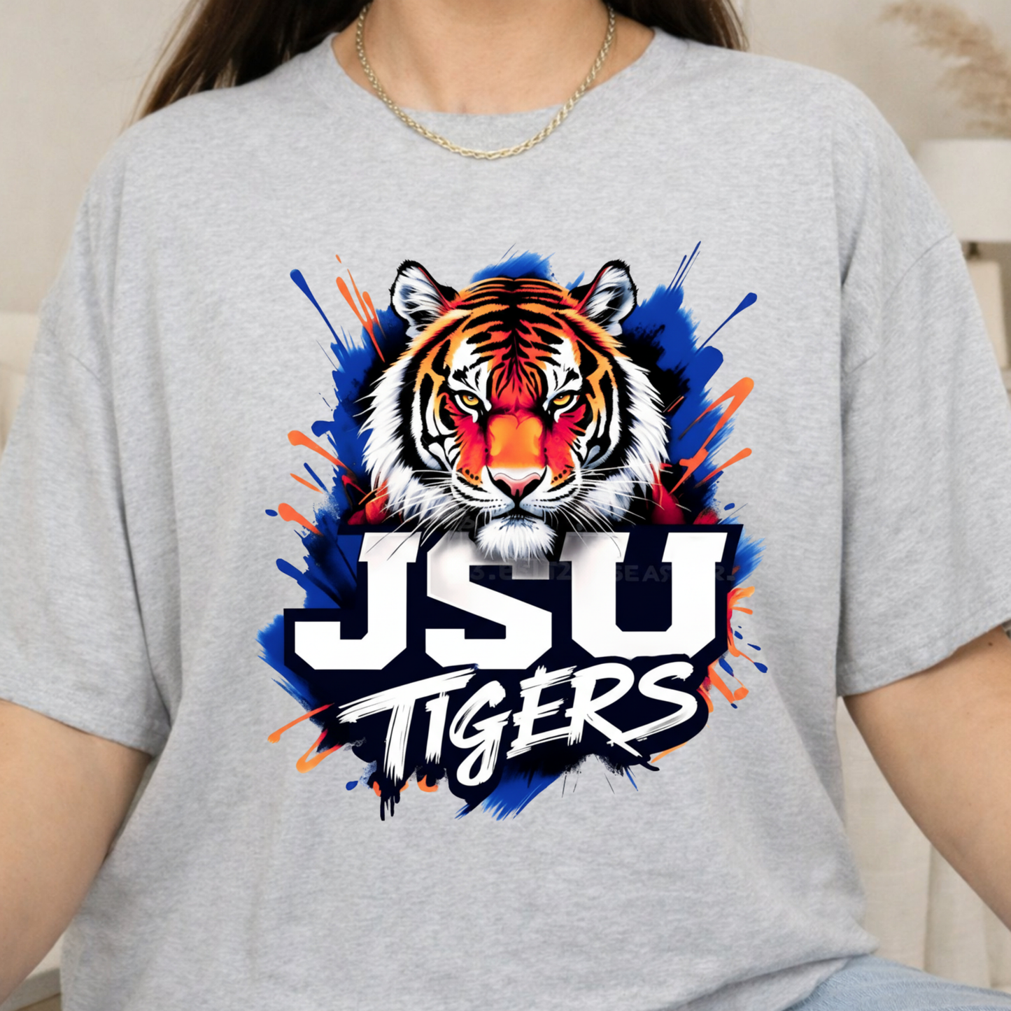 A person wearing a light heather gray t-shirt with a large graphic of a tiger's face and the text 'JSU TIGERS' in white over a blue and orange paint splatter design.