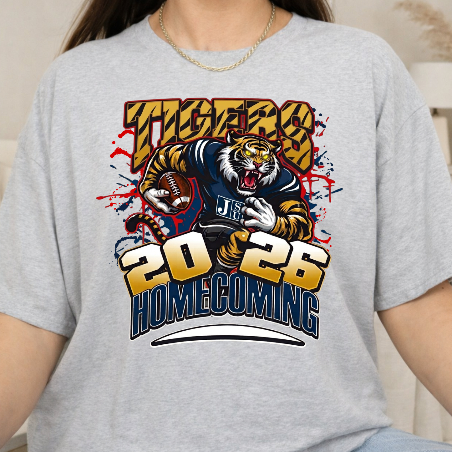 A person wearing a light heather gray t-shirt with a large graphic print featuring a tiger mascot, the word 'TIGERS', and '2026 HOMECOMING'.