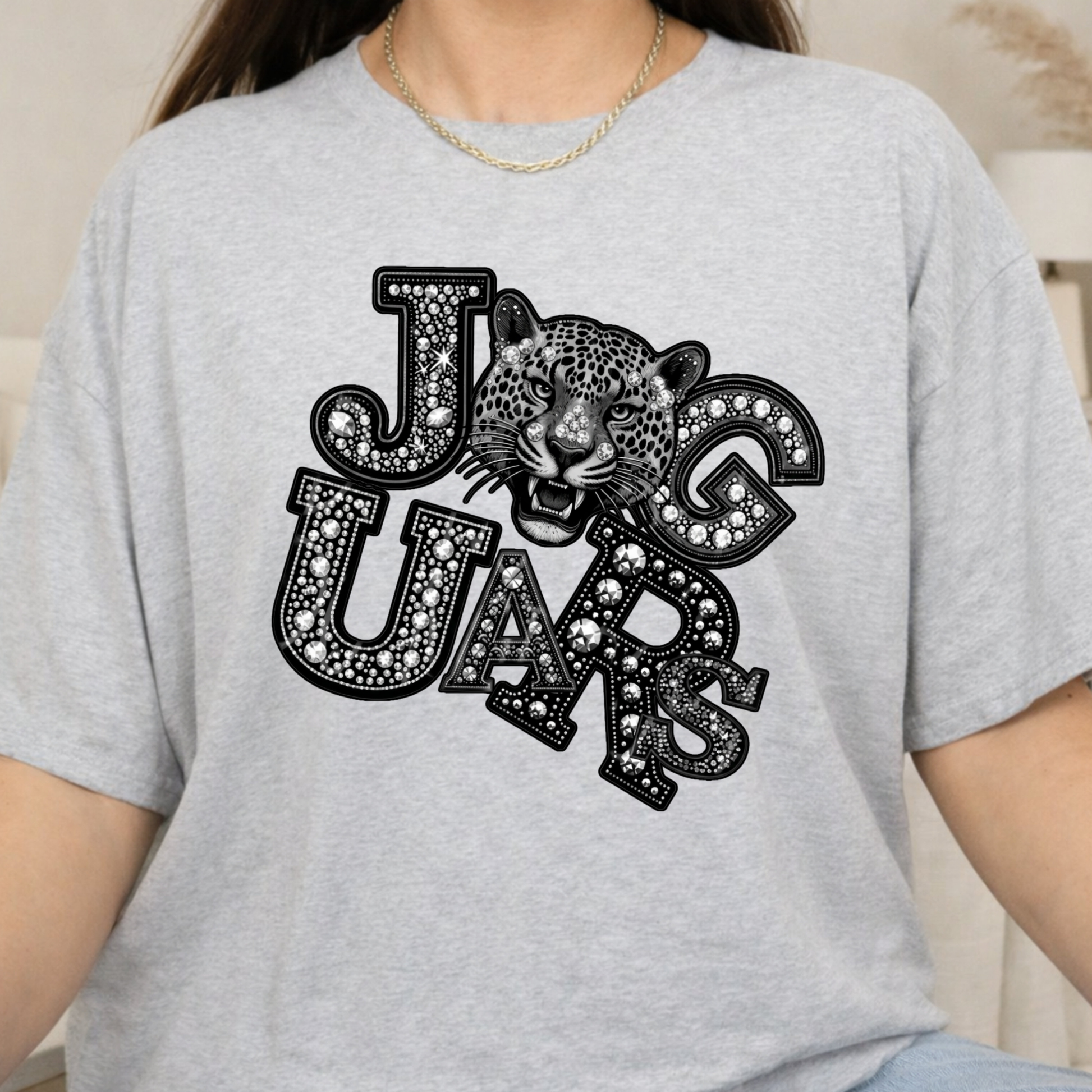 A person wearing a light heather gray short-sleeve t-shirt with a black graphic print that reads 'JUG UARS' in decorative lettering with a leopard face in the center.