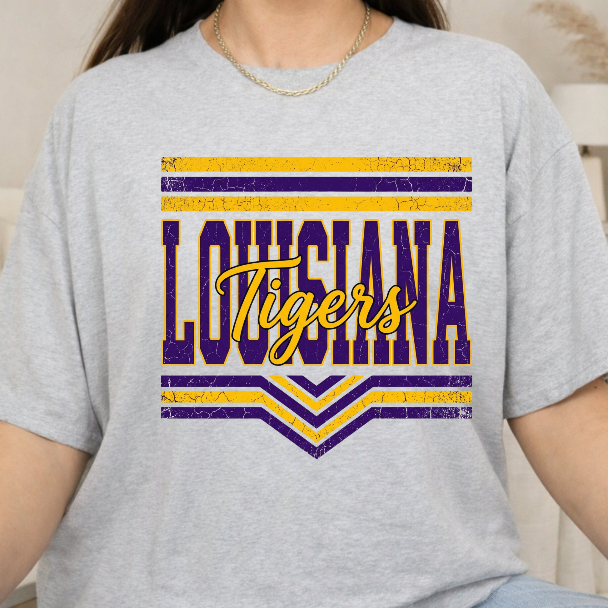 A person wearing a light heather gray graphic T-shirt with a distressed purple and yellow 'LOUISIANA Tigers' design on the front.