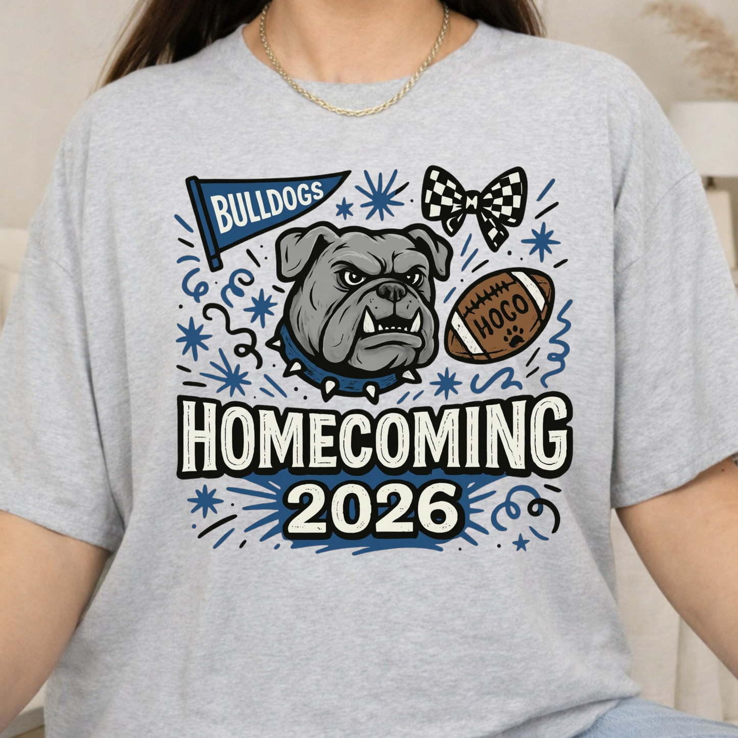 A woman wearing a light heather gray t-shirt with a graphic design that reads 'BULLDOGS HOMECOMING 2026' featuring a bulldog, football, and bowtie illustration.