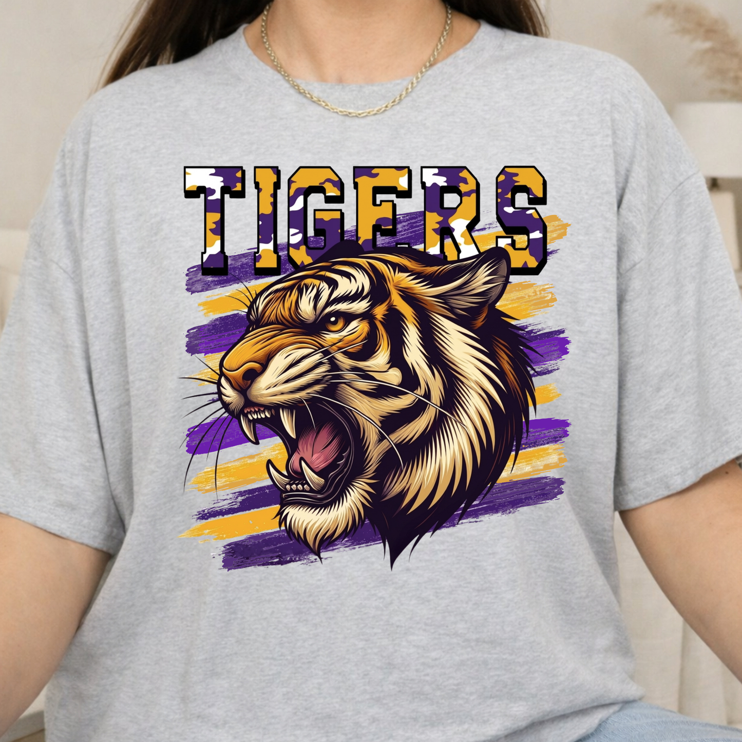 A person wearing a light heather gray T-shirt with a large graphic print featuring the word "TIGERS" in purple and gold patterned letters above a roaring tiger illustration on purple and gold brushstroke stripes.