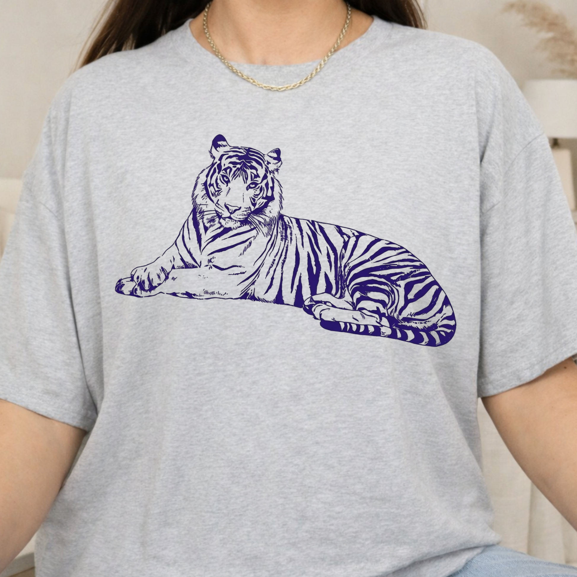 A person wearing a light heather gray short-sleeve T-shirt with a large dark blue line-art tiger graphic across the chest, photographed indoors against a soft neutral background.
