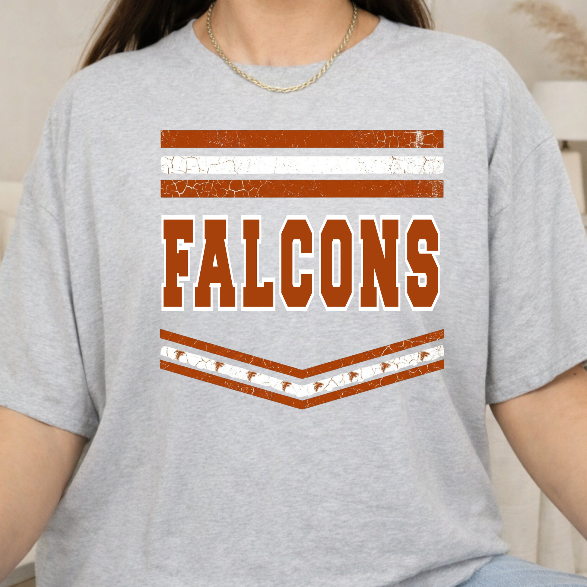 A person wearing a light heather gray short-sleeve T-shirt with a distressed collegiate-style graphic on the chest featuring brown and white stripes and the word "FALCONS" in brown block letters outlined in white.