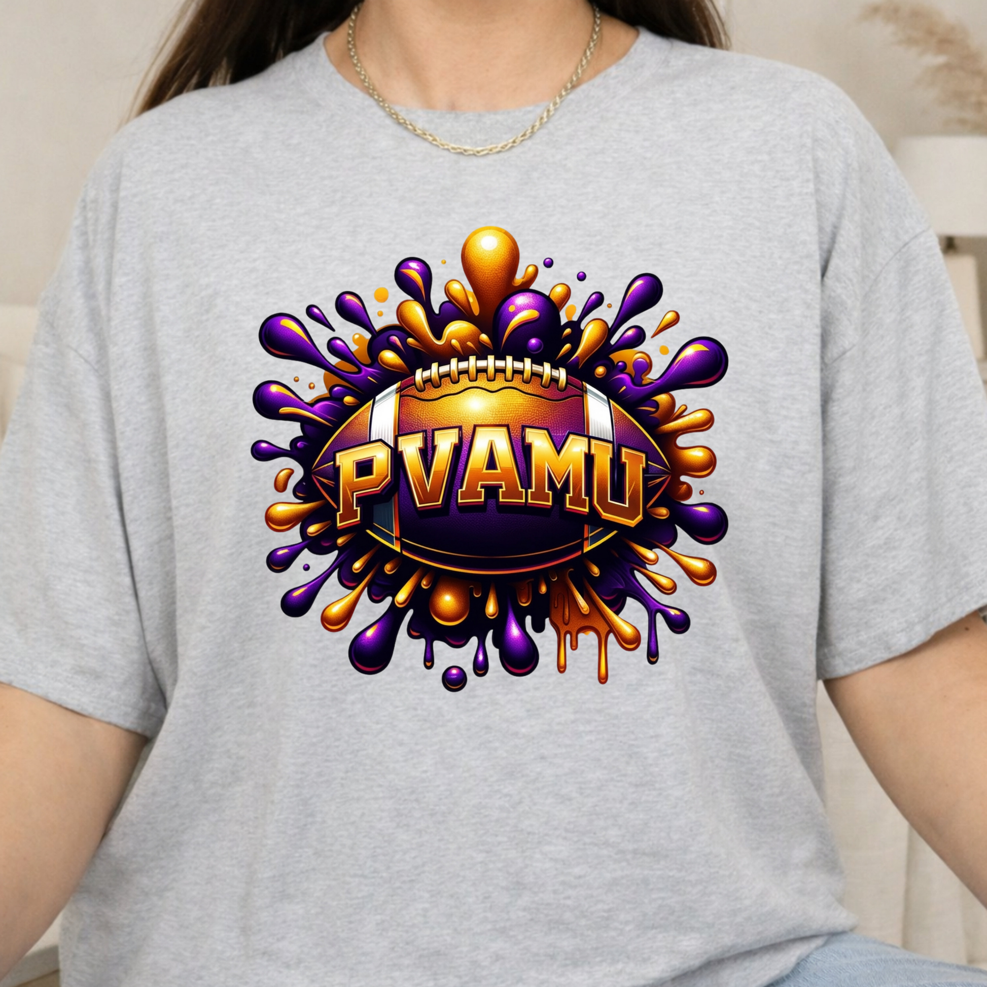 A person wearing a light heather gray t-shirt with a large graphic on the chest featuring a football and the text 'PVAMU' in gold and purple paint-splatter artwork.