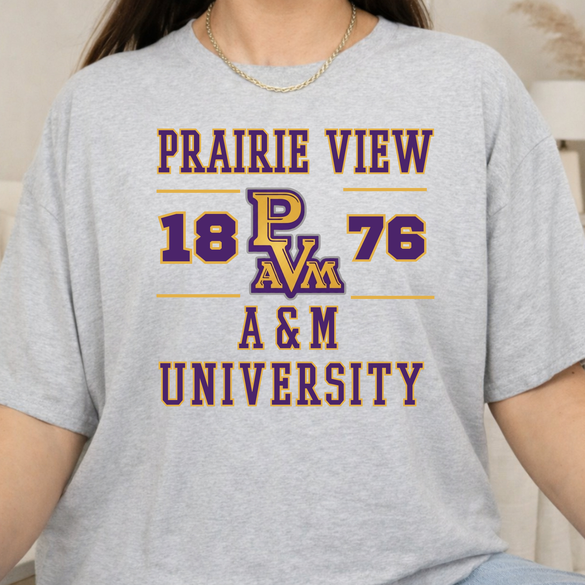 A person wearing a light heather gray graphic T-shirt with purple and gold text reading 'PRAIRIE VIEW 1876 A&M UNIVERSITY' and a stylized 'PAVM' logo on the chest.