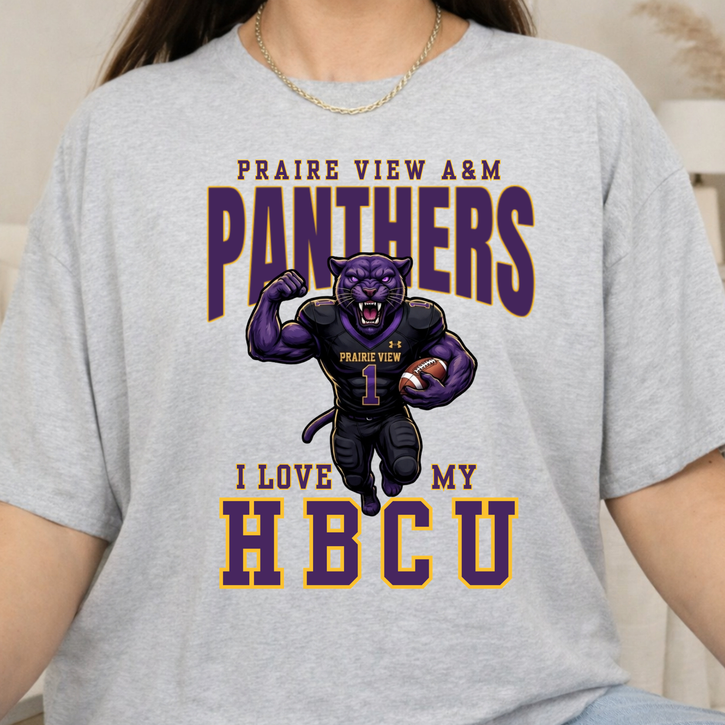 A person wearing a light heather gray graphic T-shirt. The shirt features a purple and gold print with the text 'PRAIRIE VIEW A&M PANTHERS I LOVE MY HBCU' and an illustration of a panther mascot in a football uniform.