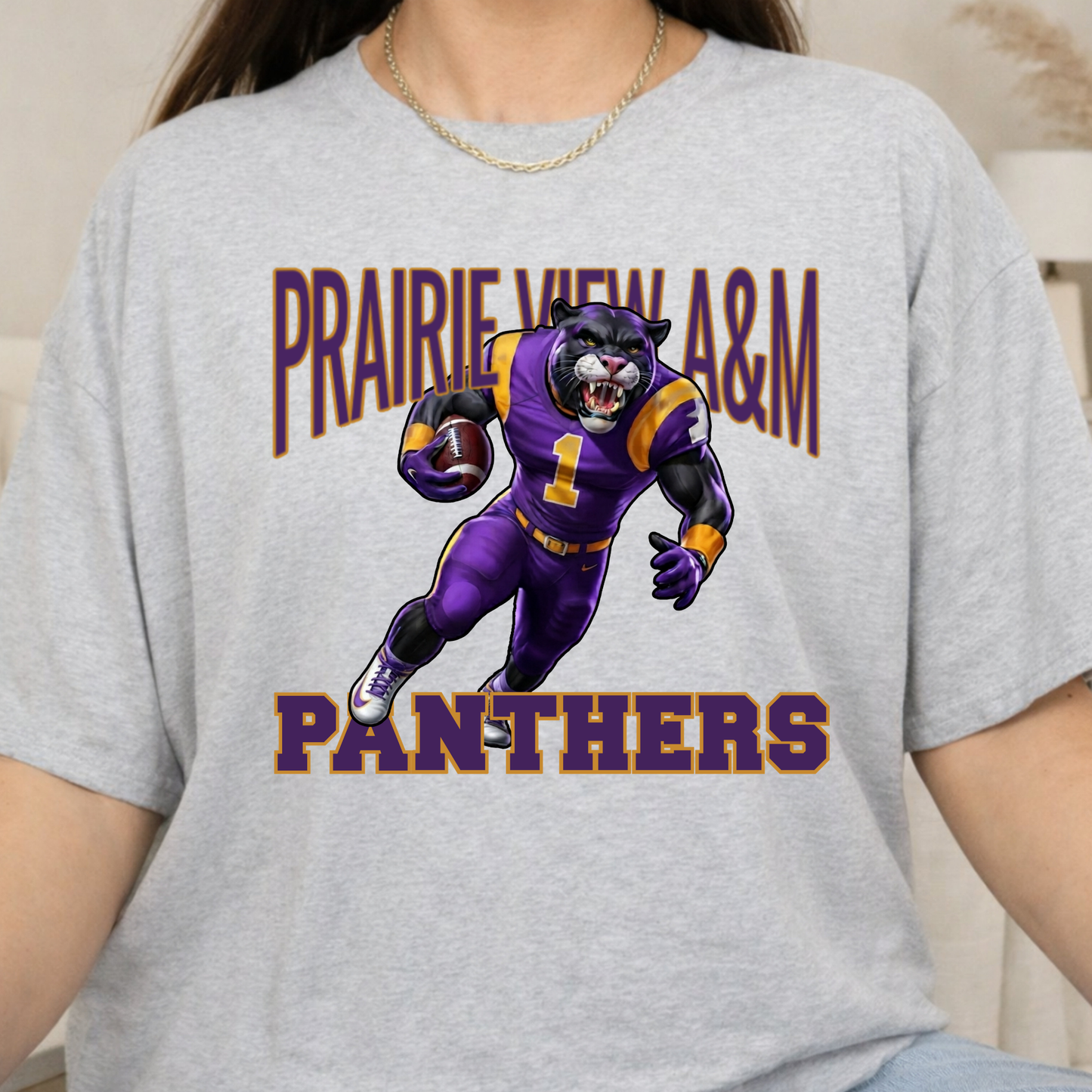 A person wearing a light heather gray graphic T-shirt featuring a purple and gold cartoon panther mascot with the text 'PRAIRIE WEE A&M PANTHERS' on the front.