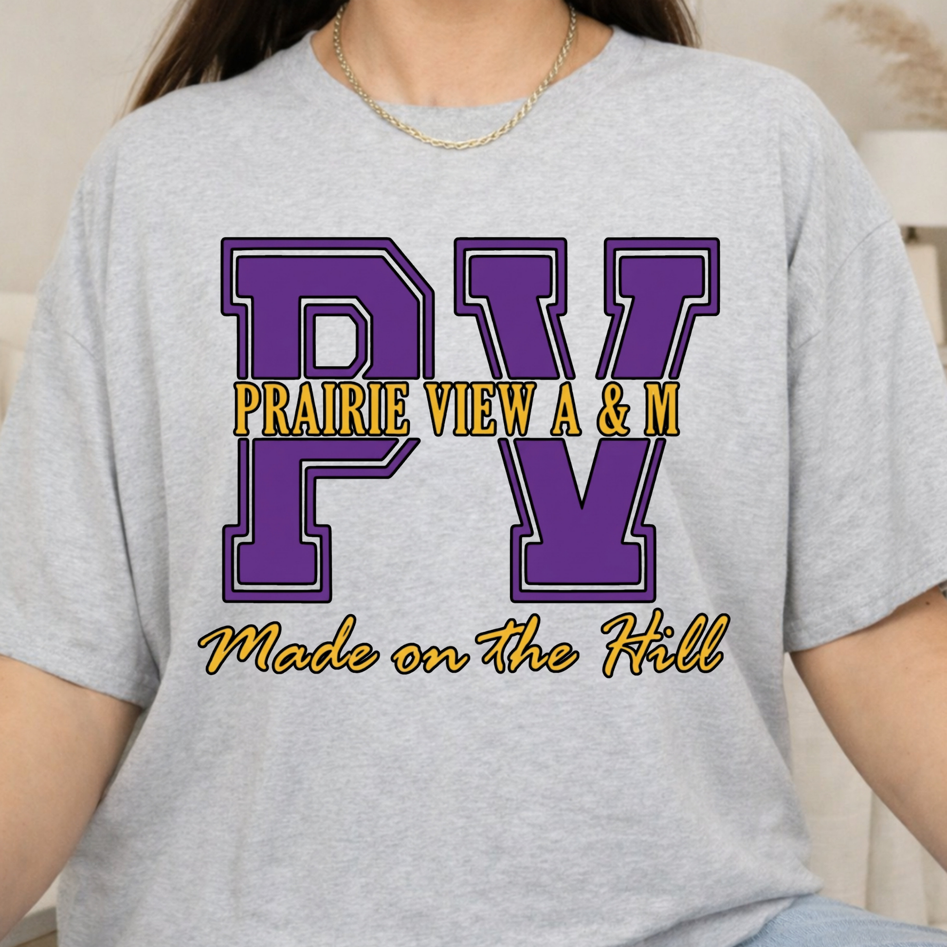 A person wearing a light heather gray graphic T-shirt with large purple 'PV' letters and yellow text reading 'PRAIRIE VIEW A & M' and 'Made on the Hill'.