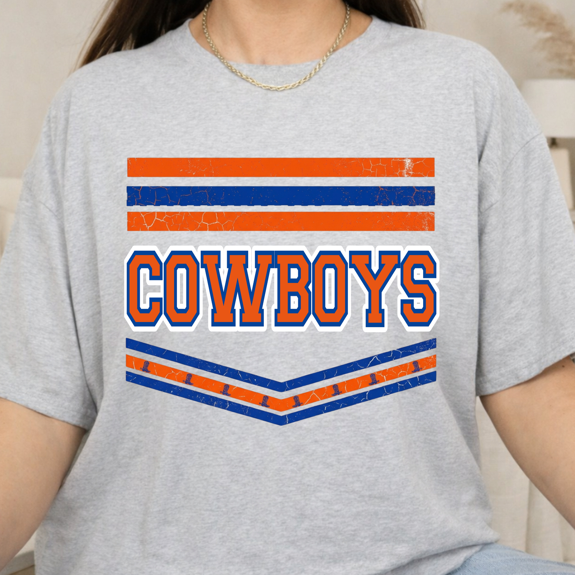 A person wearing a light heather gray t-shirt with a distressed graphic that reads 'COWBOYS' in orange and blue block letters, framed by orange and blue horizontal stripes.