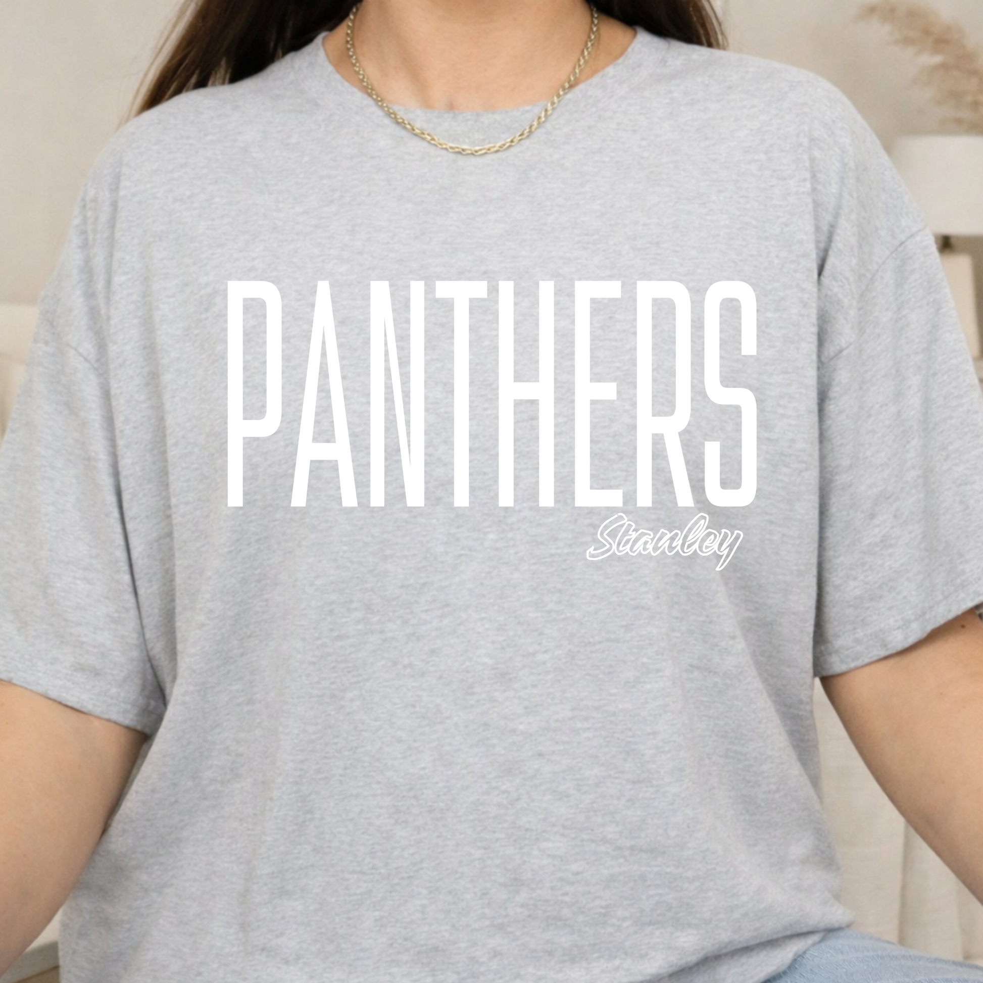 A person wearing a light heather gray short-sleeve T-shirt with large white 'PANTHERS' text and a smaller white 'Stanley' script logo on the front.