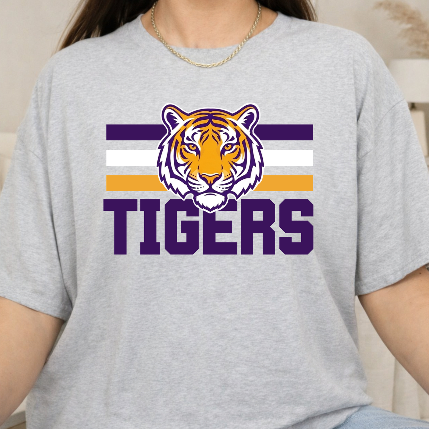 A person wearing a light heather gray t-shirt with a purple, white, and orange tiger graphic and the word 'TIGERS' printed on the front.