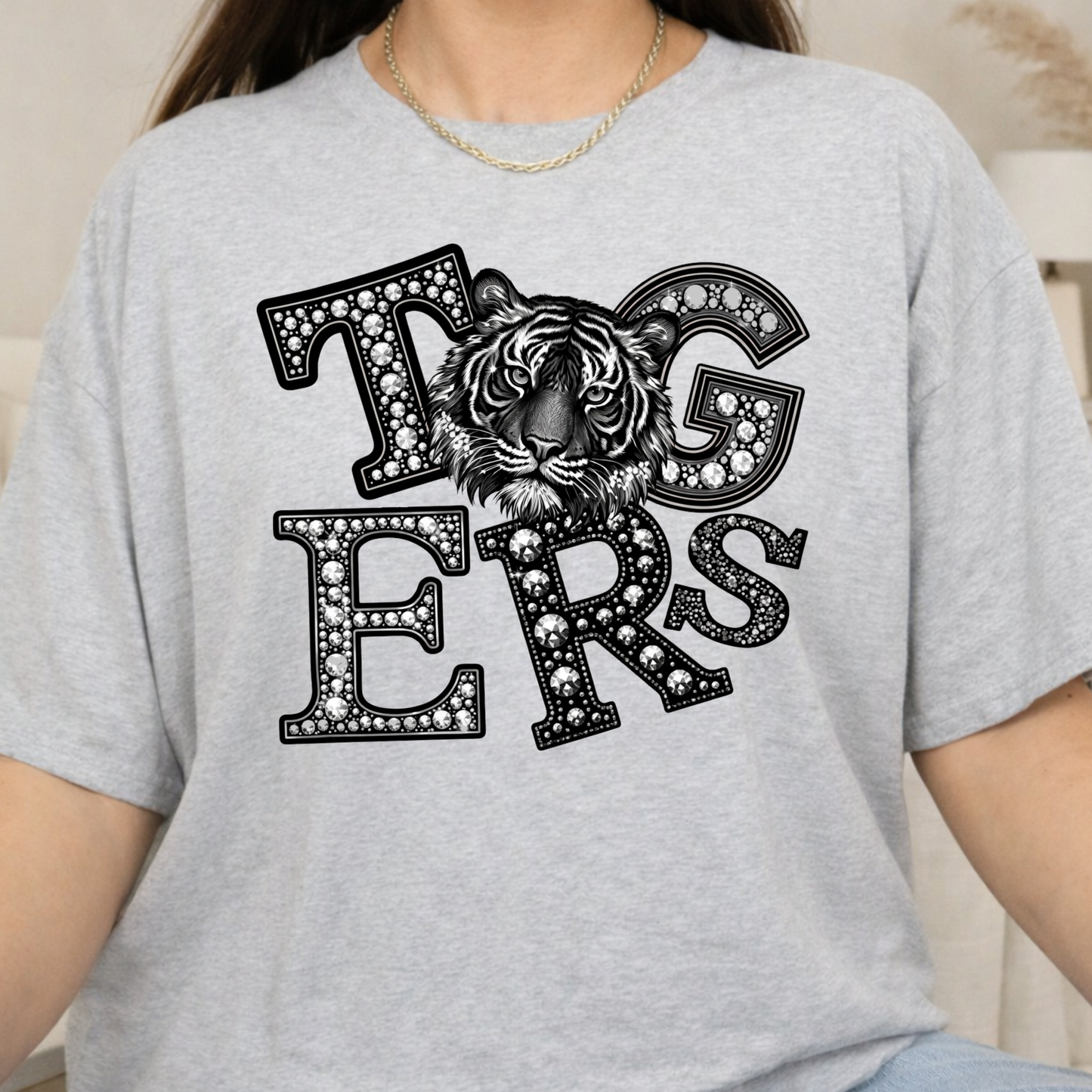 A person wearing a light heather gray short-sleeve t-shirt with a large black graphic on the chest spelling 'Tigers' in a diamond-studded font, with a tiger face integrated into the design.