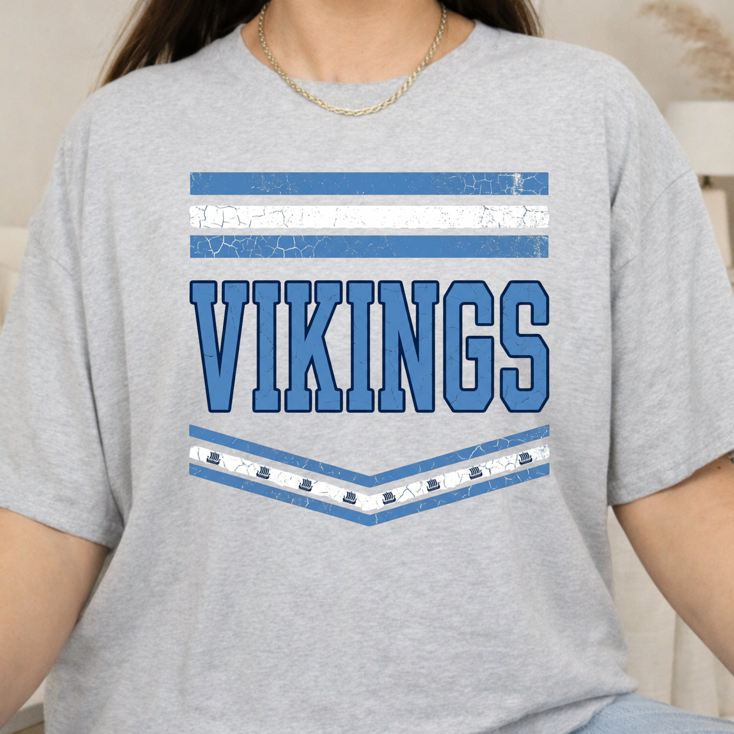 A person wearing a heather gray short-sleeve T-shirt with a distressed blue and white 'VIKINGS' graphic on the front, shown from the shoulders to the hips in a softly lit indoor setting.