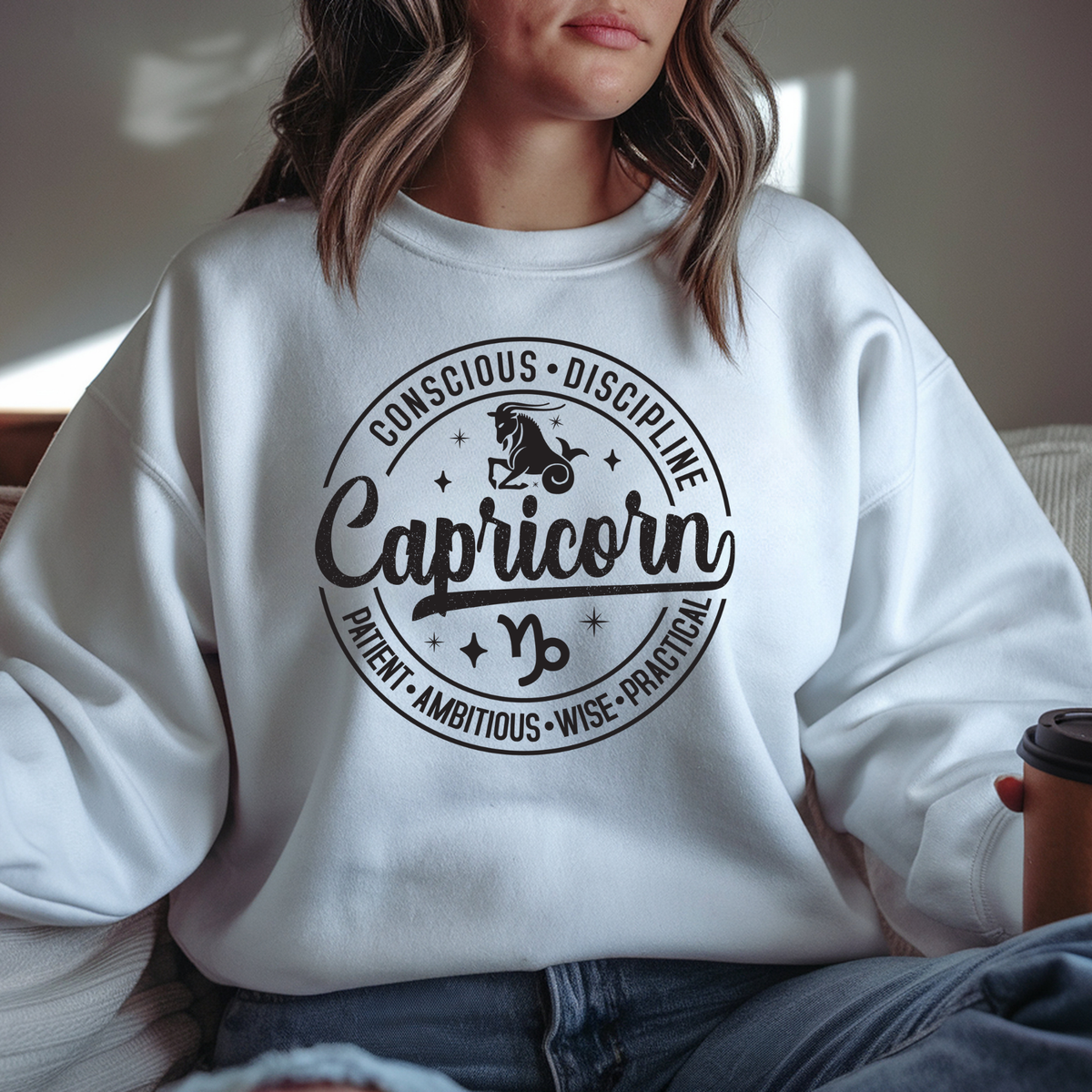 A woman wearing a white crewneck sweatshirt with a black circular graphic that reads 'Sagittarius' and lists traits like Honest, Independent, Optimistic, and Intelligent. She is sitting indoors holding a coffee cup.