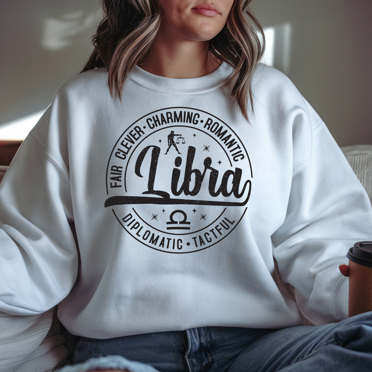 A woman wearing a white crewneck sweatshirt with a black Libra zodiac graphic on the front, sitting indoors and holding a coffee cup.