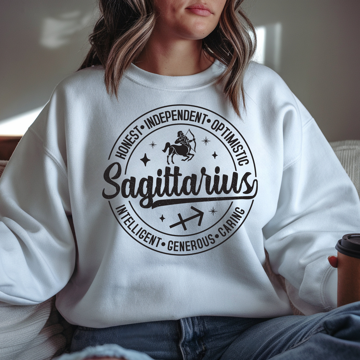 A woman wearing a white crewneck sweatshirt with a black circular graphic that reads 'Sagittarius' and lists traits like Honest, Independent, Optimistic, and Intelligent. She is sitting indoors holding a coffee cup.