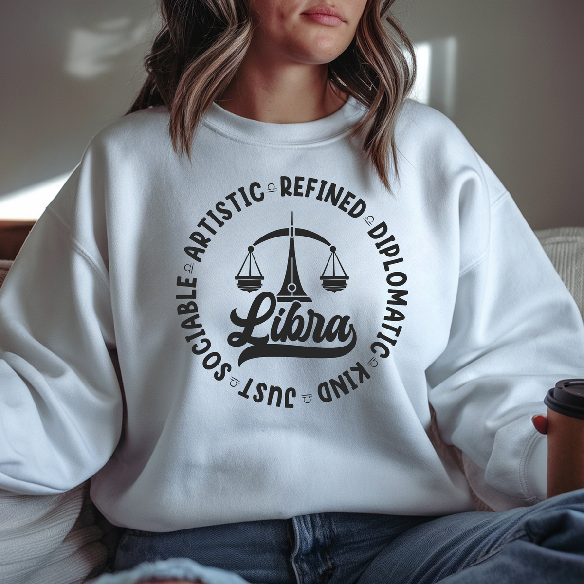 Woman wearing a white crewneck t-shirt featuring a black circular Libra zodiac graphic with a scales illustration, the word "Libra," and trait keywords like artistic, refined, diplomatic, kind, just, and sociable.