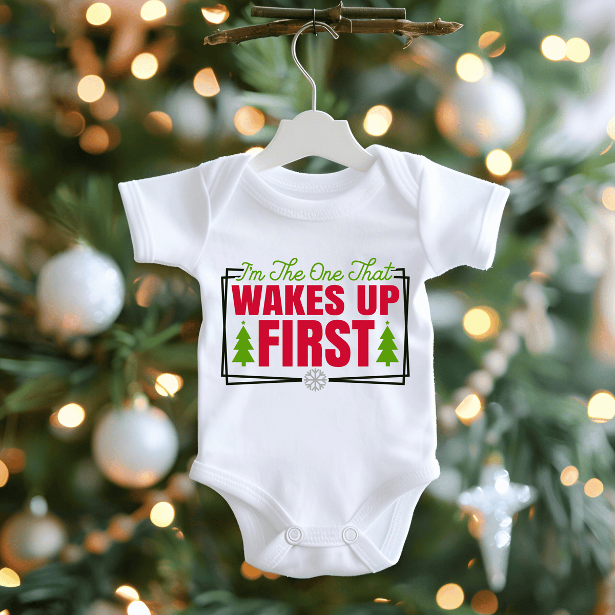 A white baby bodysuit hanging on a white hanger from a decorative branch, featuring a festive Christmas graphic that reads 'I'm The One That WAKES UP FIRST' in green and red text with two green trees. The background is a blurred Christmas tree with warm lights and silver ornaments.