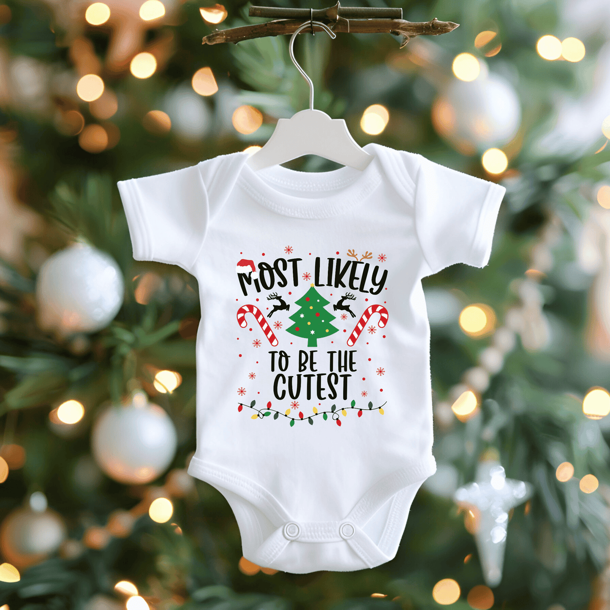 A white short-sleeve baby bodysuit hanging on a white hanger from a rustic branch, featuring a festive Christmas graphic with the text 'MOST LIKELY TO BE THE CUTEST' and illustrations of a tree, candy canes, birds, and lights. The background shows a blurred Christmas tree with warm bokeh lights.