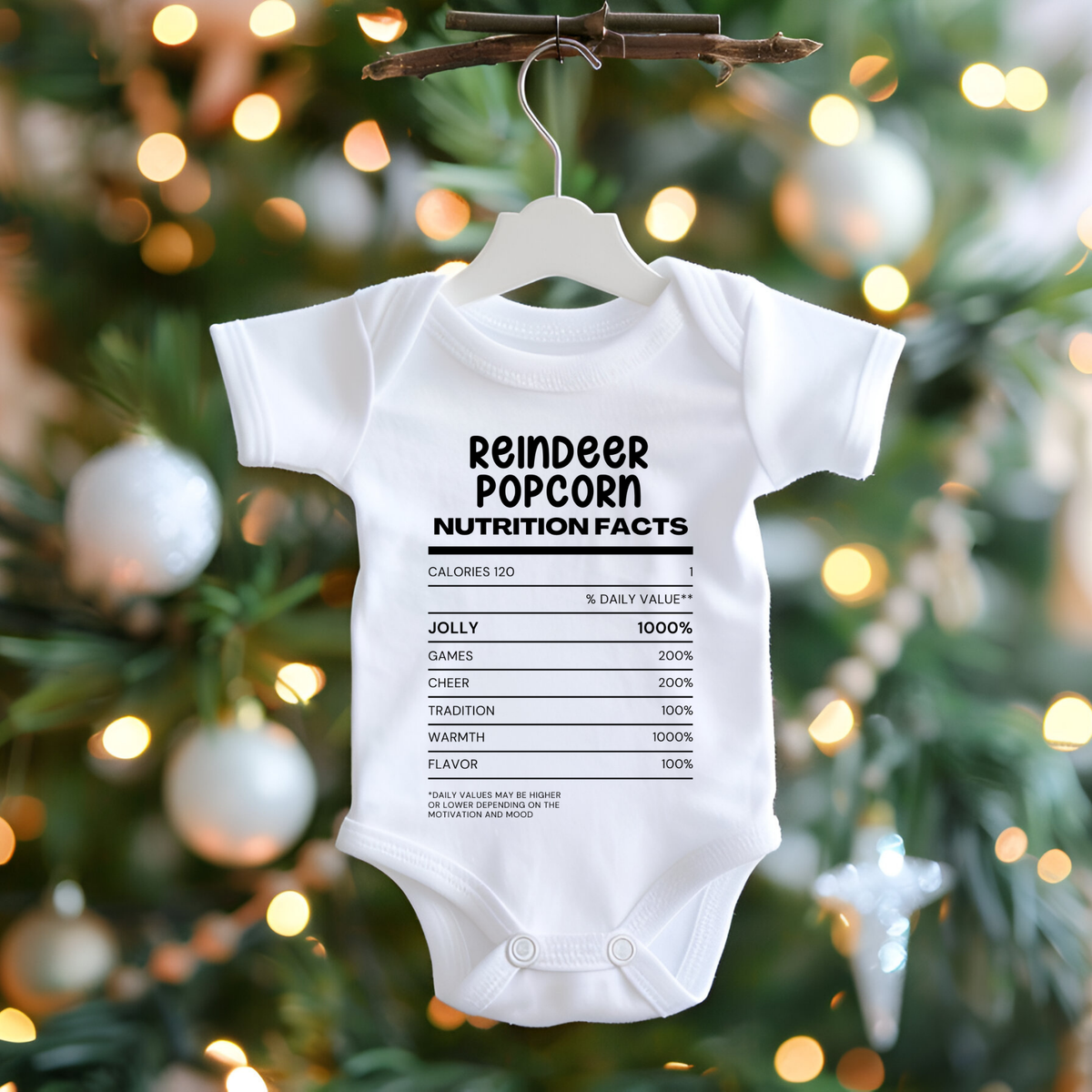 A white short-sleeve baby bodysuit hanging on a white hanger from a twig, featuring a festive Christmas graphic that reads 'Ain't No Family Like The One I Got' in green lettering with a Santa hat, snowman, reindeer, and candy cane illustrations. The background shows a blurred Christmas tree with warm lights and ornaments.